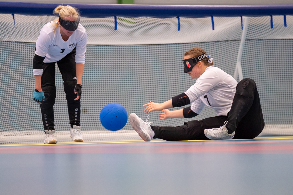 Miina Grönroos istumassa lattialla, tiputtamassa palloa vieressä seisten odottavalle Raisa Kuuselalle.